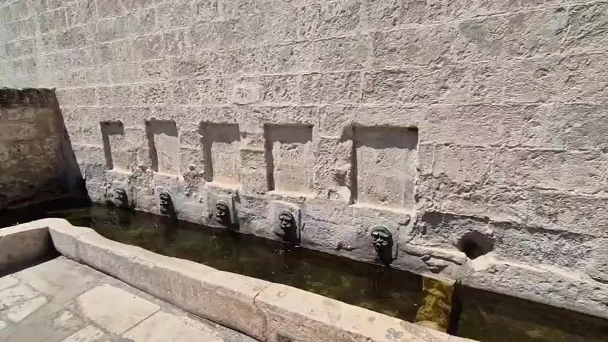 The video shows a public fountain with a long stone basin. The basin is filled with water and has several decorative lion heads that appear to be spouts, from which water is flowing. The fountain is built against a large stone wall, and there are arched structures behind the fountain that also seem to be part of the water system. The area around the fountain is paved with stone tiles. The environment appears to be outdoors, with sunlight casting shadows, and buildings visible in the background. The footage pans across the fountain and its surroundings, offering different perspectives of the structure and the water.