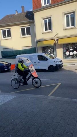 A man in a neon yellow jacket and helmet rides a black tricycle, with a clear dome windscreen, down a European street on a sunny day. The tricycle has a basket on the back. The street has buildings, parked cars, and a bus driving in the opposite direction. The man is riding with traffic. Other parked bicycles are present.