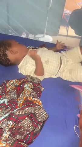 A baby is lying on a blue bed, on their back. The baby is wearing a light-colored onesie with a checkered pattern and a small graphic print. To the left of the baby is a vibrantly patterned blanket with red, orange, and black designs. In the background, a white standing fan is visible. The baby's left hand is in their mouth. The baby moves their right hand near their face. The baby then turns their head and looks towards the camera.