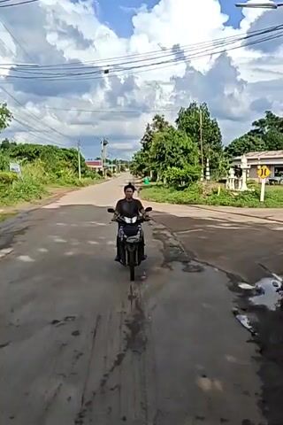 A person is riding a motorcycle down a paved road. The person is wearing a dark shirt and dark pants. The motorcycle is black and silver. The road is lined with trees and grass. There are power lines overhead. The sky is cloudy. The video is shot from a low angle, following the motorcycle.