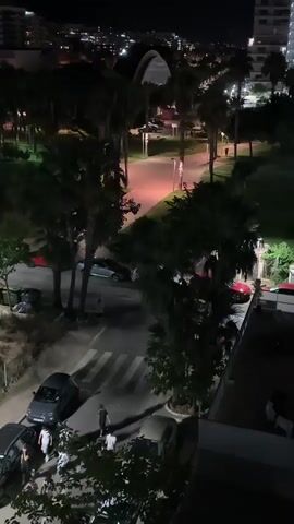 This media file is a nighttime aerial shot of a street scene in Les Amplaries, Spain. The primary subjects are several vehicles, including cars and what appears to be a motorcycle, parked and driving on the road and in adjacent parking areas. Palm trees are a prominent feature of the landscape, casting shadows across the scene. A few people are visible, some standing on the sidewalk near parked cars, and one walking across a crosswalk.

The activity appears to be the normal flow of nighttime street life. Cars are parked along the street and on the side, and one car is in motion. People are gathered on the sidewalk, suggesting a social or residential area. The lighting is artificial, provided by streetlights and lights from buildings, creating a typical urban nighttime ambiance. The overall impression is one of a quiet evening in a Mediterranean coastal town.

Notable details include the time of day, which is clearly night due to the dark sky and artificial lighting. There are no obvious cues about the weather, but the scene is well-lit and appears clear. The presence of palm trees and the style of buildings in the background suggest a warm climate. The general atmosphere is peaceful and ordinary. There is no visible text in the media file.