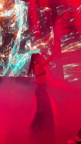 A woman in a sparkly two-piece outfit dances center stage. The background is a large screen displaying a colorful, abstract, cosmic-like image. Stage lighting casts a purple and blue glow, with smoke machines adding to the atmosphere. A DJ booth is visible to the left of the stage.