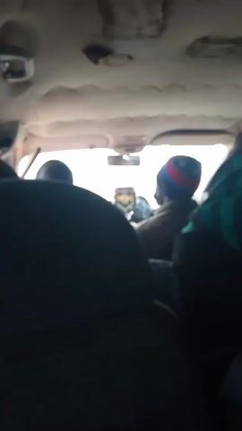 The video shows a view from the inside of a moving vehicle, likely a car or van. The perspective is from the back seat, looking towards the front. The front passenger seat is visible, with a person wearing a hat. The focus shifts to the side window, revealing a landscape of dry, open land with sparse vegetation. In the distance, there's a hint of hills or mountains under a pale sky. The vehicle's interior includes a seatbelt and the headliner.