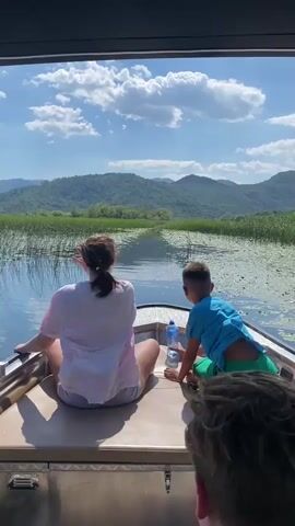 A wide shot captures two young people from behind, a woman and a boy, sitting in the back of a motorboat that is moving forward through a body of water filled with lily pads. The woman is wearing a pink and white shirt and grey shorts. The boy is wearing a blue shirt and green shorts. A bottle of water sits between them. In the background, lush green hills rise under a bright blue sky dotted with white clouds. The boat appears to be moving along a channel with tall green reeds and lily pads on either side. A third person's hair is visible in the foreground.