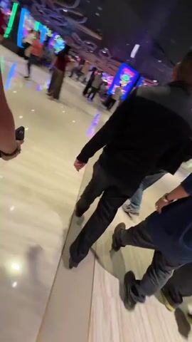 The camera moves through a casino with a white, marble-tiled floor and a high, dark ceiling with exposed ductwork. The floor is divided into sections by narrow, inset stripes, some of which are illuminated with blue or purple light. Several people are visible, mostly from the back, standing near slot machines. Some people are walking in different directions. The slot machines are brightly lit, with screens showing colorful graphics. The overall impression is of a crowded, active gambling environment. The camera angle is low and slightly tilted, as if held by a person walking through the casino.
