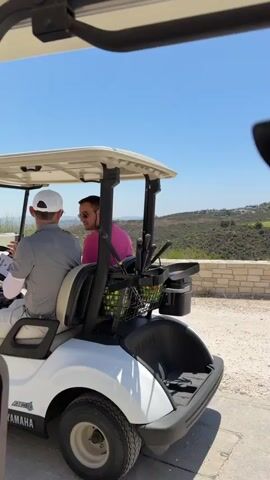The video shows two men sitting in a white Yamaha golf cart. The man in the driver's seat is wearing a gray polo shirt, a white cap, and light-colored pants. He has on white arm sleeves and white athletic shoes. The man in the passenger seat is wearing a bright pink shirt and dark shorts. He has sunglasses on and appears to be talking to the driver. The golf cart is parked on a paved surface with a scenic, hilly landscape in the background. The sky is clear and blue. Golf clubs are visible in the back of the golf cart. There is a basket on the floor of the golf cart, and a sign with the number 56 is on the side. The lighting suggests it is daytime.