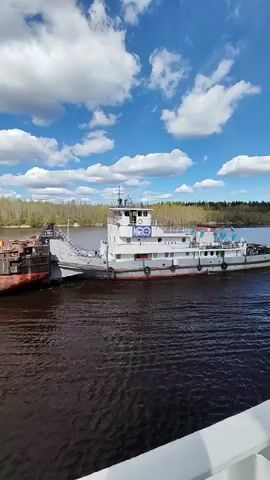 The video shows a white boat sailing on a river under a partly cloudy sky. The boat has multiple levels and a bridge on top. It is connected to another smaller vessel by a ramp. The river water is dark, and the banks are lined with trees.