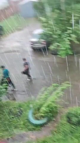 A person wearing dark clothing and a light-colored shirt runs across a wet, paved area, appearing to be in a residential courtyard during a heavy rain shower. Two cars, one black and one beige, are parked in the background. Lush green foliage is visible on the left side of the frame and in a repurposed tire planter on the right. The rain is intense, creating visible streaks and puddles on the ground.
