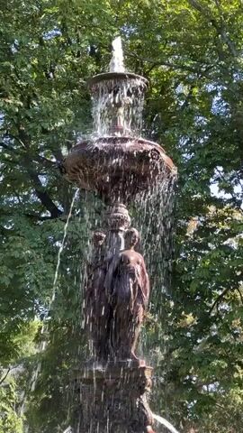 A bronze fountain featuring three female figures stands in the center of the frame. The figures are clustered together, with water cascading down from a large, ornate basin above them. The fountain is surrounded by lush green trees, suggesting an outdoor setting, possibly a park or garden. Sunlight dapples through the leaves, illuminating the water and the bronze sculptures. At the bottom of the frame, the base of the fountain is visible, with more water flowing into a pool. In the lower left corner of the frame, two individuals are partially visible, standing near the pool. One individual appears to be wearing a light-colored shirt and dark pants, while the other's attire is less distinct. The water is in constant motion, creating a dynamic and refreshing visual.