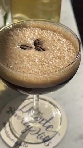 The video shows a close-up of an espresso martini sitting on a coaster that says "Polo Lounge Beverly Hills," adorned with three coffee beans as garnish. In the background, a glass carafe of a light-colored beverage and white flowers are seen. As the camera zooms out, the location is revealed to be an outdoor patio setting, adjacent to a swimming pool area with several individuals sunbathing. The scene is open to the elements, with sunlight streaming in and casting shadows on the wooden deck and patio furniture.