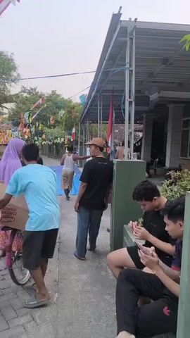 The video shows an outdoor scene with several people and decorations. In the foreground, two young men are sitting and looking at their phones. Behind them, a man in a blue shirt and shorts walks away from the camera, followed by another man in a black shirt and a brown cap. To the left, a woman in a purple headscarf and a patterned dress is standing with a young girl in an orange shirt and black pants. A pink bicycle is positioned next to them. Further back, an Indonesian flag is flying, and there are various colorful banners and decorations strung up. The environment appears to be a residential area, possibly a village, with some houses visible and trees in the background. Some bags or packages are hanging from strings in the decorated area. The overall atmosphere seems to be a casual gathering or celebration.
