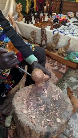 A person is shown sitting and carving a small wooden sphere with a tool. The person is wearing a colorful, patterned jacket. They sit in front of a tree stump covered with wood shavings, which serves as their workspace. Surrounding the artisan are various completed wooden carvings, including elephants, figures, and other animal shapes, displayed on a table behind them. The scene takes place in an indoor market or workshop setting with brick flooring.