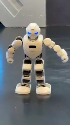 A small white humanoid robot dances in place on a black floor. The robot has a rounded head with blue glowing eyes, a white torso with a logo, and articulated arms and legs with black and white components. The robot's arms move in a rhythmic fashion, and music plays in the background. The environment is a room with a gray-and-white wall and some other indistinct objects.