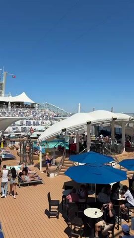 The video captures a lively outdoor scene on a sunny day, likely on a cruise ship or resort deck. In the foreground, several people are seated at tables under blue umbrellas, some appearing to be dining or socializing. The deck is made of light-colored wood. In the mid-ground, a swimming pool is visible, with people lounging on chairs and walking around it. Further back, a tiered seating area, reminiscent of an amphitheater, is set up. A large white canopy covers part of the area, providing shade. The sky is a clear, bright blue. The overall impression is one of leisure and recreation.