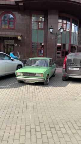The video shows a light green Lada sedan parked on a paved surface. The car is facing the camera. The license plate reads "E060 AT 977 RUS." The car is parked next to a brown Lada station wagon and a white scooter. A person wearing a blue jacket is visible on the scooter. A building with multiple windows is in the background. There is a lamppost with two lights to the right of the green Lada. The camera pans slightly around the front of the green car.