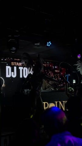 The video shows a nightclub scene. In the background, there's a brightly lit display that says "STAR ON STAGE DJ TONI" above a cityscape image. In front of the display is a DJ booth where a DJ wearing a leopard print shirt and a dark cap is working. Two performers in glittery costumes with large feather wings are dancing on either side of the DJ booth. Patrons are visible in the foreground, some holding up phones to record the performance. The name "POINT" is illuminated on the front of the DJ booth. Lighting fixtures above cast colored spotlights, creating a dynamic and lively atmosphere.