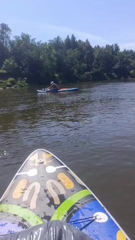 The video shows a first-person perspective of someone on a paddleboard on a river. Another person is visible on a separate paddleboard in the distance, further up the river. The water is calm, and the riverbanks are lined with trees. The sky is clear and blue. The person in the foreground is looking forward towards the other paddleboarder.