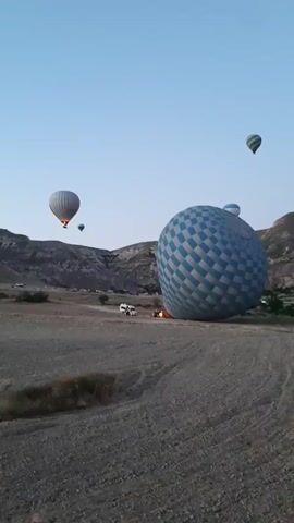 A hot air balloon with a blue and white checkered pattern is deflated on the ground in a dry, rocky field. Several other hot air balloons are inflated and ascending into the sky, scattered across a vast landscape with rolling hills and rock formations. The sky is clear and light blue, suggesting an early morning or late evening time. In the background, there are a few vehicles and people near the deflated balloon, possibly preparing for or packing up after a flight. The overall scene depicts a hot air balloon activity in a natural, rugged environment.