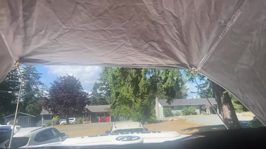The video is taken from inside a tent, looking out through an opening. Outside, there is a residential area with houses and trees under a partly cloudy sky. Several cars are parked in front of the houses. The tent is made of a light-colored fabric and is supported by poles, which are visible as metal rings where the fabric is attached to the poles. The sun is shining brightly, casting shadows on the ground. The overall impression is of a sunny day in a suburban environment.