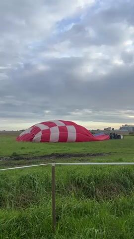 The video shows a large, deflated hot air balloon lying on its side in a grassy field under a cloudy sky. The balloon has a red and white checkered pattern. Several people are standing near the rear of the balloon, some appearing to be involved in packing it up. A vehicle is also visible behind them. The foreground is dominated by tall green grass, with a wooden post and a wire fence running horizontally across the image. The background shows a flat, open landscape with distant trees or buildings. The overall impression is one of the end of a hot air balloon flight or preparation for transport.