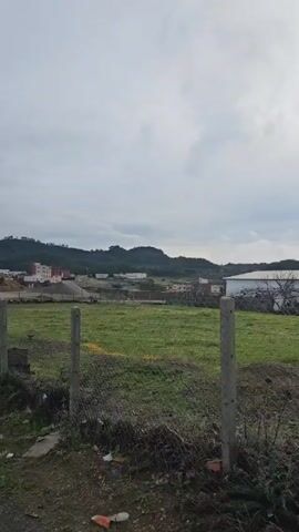The video shows a rural landscape on a cloudy day. A chain-link fence runs along the side of a narrow, wet road. A field of green grass stretches beyond the fence, dotted with some small white flowers. In the distance, there are hills covered with sparse vegetation. Several buildings are visible scattered in the landscape, including some with red roofs. The camera slowly pans to the right, revealing more of the road and the landscape. There are puddles and streams of water running along the road, indicating recent rain. The sky is overcast, and the sun is diffused through the clouds.