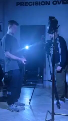 The video shows two men in a room that is illuminated by a bright blue light. The room has the words "PRECISION OVER" displayed on the wall. One man is on the left side of the frame, wearing a gray t-shirt and dark pants with white stripes. He is holding a small, dark object in his hands and looking down at it. The second man is standing behind a camera mounted on a tripod. He is wearing a dark jacket with white stripes and khaki pants. The camera is pointed towards the first man. There is a bright light attached to the top of the camera. The second man appears to be adjusting or operating the camera.