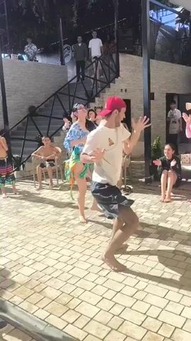 A group of people are gathered outdoors on a paved patio. In the foreground, a man in a white t-shirt and black shorts, wearing a red baseball cap, is dancing. To his left, a woman in a blue and white patterned top and a green and orange skirt is also dancing. Several other people are seated and standing around watching them. Some people are on a raised platform in the background, which has a set of stairs leading up to it. The environment appears to be a resort or entertainment venue with some vegetation visible in the background.
