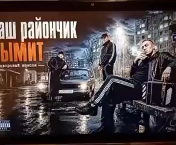 A computer screen displays album art for a Russian rap song titled "Nash Rayonchik Dymit," which translates to "Our Neighborhood is Smoking." The art features three men dressed in tracksuits in a dimly lit, urban setting with a classic car and apartment buildings. The man in the foreground, seated on a bench, is smoking and playing an accordion. The man in the middle stands near the car, and the man on the left leans against it. The overall mood is gritty and atmospheric, with wet streets reflecting the streetlights, suggesting a nighttime scene. There is a parental advisory label in the lower left corner. The video appears to be a short clip, possibly a preview or advertisement for the song, with the audio featuring the beginning of the track.