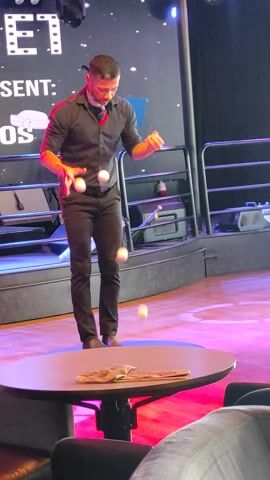 A male performer is juggling three white balls on a stage. He is wearing a black shirt with a red scarf tied around his neck, dark pants, and dark shoes. The performer is in mid-air, with one ball above his head, one in his left hand, and another in his right hand. He is illuminated by stage lights, casting a vibrant pink and purple glow on the stage floor. In the background, there is a black curtain, metal railings, and a backdrop with white lettering that reads "PRESENT:" and a white hat symbol. There are also several spotlights visible. To the right of the performer, there is a round table with some objects on it, partially obscured by a person's arm and shoulder in the foreground. The overall atmosphere appears to be a live performance or show. The lighting suggests it is evening or indoors.