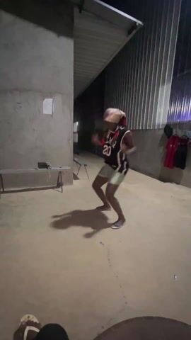 A person dances alone in an outdoor space with concrete floors and walls. They wear a head covering, a dark tank top, light-colored shorts, and sandals. A metal bench and a small electronic device rest against one wall. Metal corrugated sheeting forms the ceiling and part of another wall, where several articles of clothing hang. The lighting is dim, and the person's shadow is visible on the floor. The video is shot from a low angle, and the feet of another person are visible in the foreground.