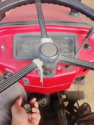 This video shows a close-up view of the dashboard of a red tractor. A person's left hand is visible operating a lever with a black knob on the left side of the steering column. The lever is moved from left to right. The tractor's steering wheel, dashboard instruments, and some of the red bodywork are visible. The background appears to be a dirt or gravel surface.