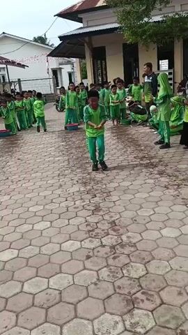 The video shows an outdoor scene, likely a schoolyard, with children in green uniforms participating in an activity. The ground is paved with hexagonal tiles, some of which are wet. In the foreground, a young boy in a green tracksuit and a black cap is the central focus. He is bending down, holding water in his cupped hands and about to pour it into a glass bottle placed on the ground. Further back, other children in similar green uniforms are lined up, watching the activity. Some adults are also present, observing the children. The environment appears to be a sunny day with a building visible in the background. The overall atmosphere suggests a playful or competitive school event.