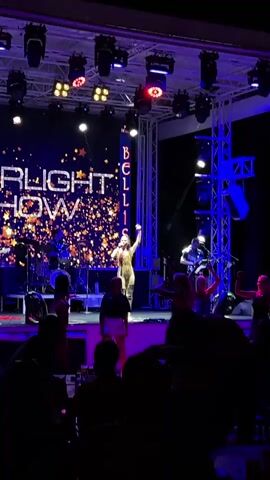 A live musical performance is taking place on an outdoor stage at night. The stage is lit with blue and purple lights, and a large screen behind the performers displays the words "STARLIGHT SHOW" with a sparkly graphic. A female singer in a sequined outfit is center stage, holding a microphone. A band, including a drummer and guitarist, is also visible on stage. Several audience members are visible in the foreground, silhouetted against the stage lights, some of whom are standing and appear to be dancing. The atmosphere appears to be festive and energetic.