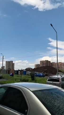 The video pans across an outdoor scene. The sky is blue with scattered clouds. In the foreground, the rear of a silver car is visible. Beyond the car, several other vehicles are parked. Further back, construction appears to be underway, with piles of dirt and construction equipment visible, along with several multi-story buildings in the background. The scene transitions to a gray, overcast sky with a light rain falling, making the road wet. More buildings and construction materials are visible. The video ends with a shot of a dark, cloudy sky over a street with a black car parked.