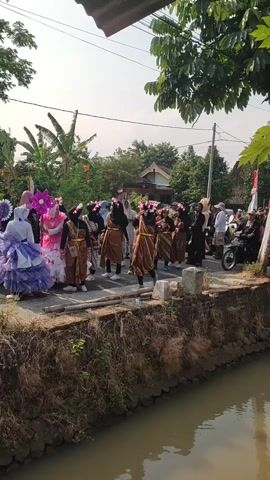 A group of people, mostly women wearing traditional clothing and flower crowns, are walking in a procession along a dirt road next to a canal. The procession includes individuals in elaborate costumes, some carrying props. There are spectators on the side of the road, and a motorcycle is parked nearby. The environment appears to be a rural area with trees and houses in the background, under a bright sky.