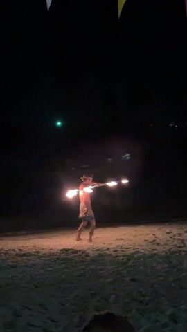 A man performs a fire dance on a beach at night. He is shirtless and wearing a skirt or loincloth-like garment. He twirls a long pole with burning flames at each end. The beach is sandy, and there are dark waves in the background. A few colored pennants are visible above. A green light is visible above the waves.
