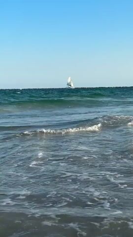 A sailboat with white sails is visible on the horizon of a deep blue sea under a clear blue sky. Waves are breaking on a beach in the foreground, with white foam visible on the crests and receding water. The camera is positioned low, facing the sea and slightly angled towards the right.