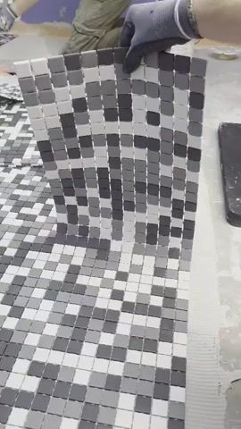 A person wearing gray gloves is in the process of tiling a floor. The tiles are small and square, arranged in a checkerboard pattern of varying shades of gray and white. The person is holding a sheet of these tiles, which is bent into a curved shape. The sheet is being placed onto a larger tiled area on the floor, which appears to be partially completed. In the background, other sheets of tiles are visible, along with buckets and tools associated with tiling work. The environment suggests an indoor construction or renovation setting.
