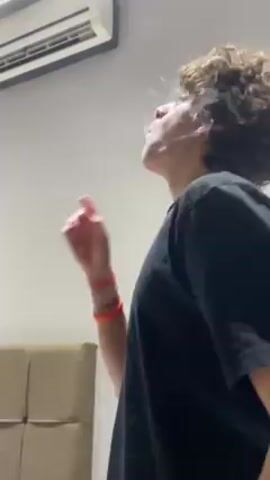 A young person with curly hair wearing a black t-shirt stands in a room with a beige wall, looking up and blowing a smoke ring towards the ceiling. An air conditioning unit is mounted on the wall above them. The person wears bracelets on their left wrist. A beige headboard is visible behind them.