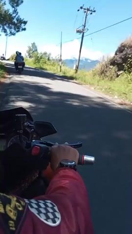 The video shows a first-person perspective of someone riding a motorcycle on a paved road, likely in a hilly or mountainous region. The road is asphalt and appears to be in good condition. To the right of the road, there is a grassy embankment with some bushes and trees, leading up to a distant mountainous landscape with scattered clouds. A utility pole with power lines runs alongside the road on the right. On the left side of the road, there are more trees and a slight incline. In the distance, on the left side of the road, a black SUV is visible, and further ahead, a motorcycle with two riders is also seen. The sky is clear and blue. The rider's hand, wearing a red long-sleeved shirt and a watch, is visible on the handlebar of the motorcycle. The shadow of the motorcycle and rider falls on the road, indicating it is daytime and the sun is out.