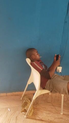 A young person, likely male, with dark skin and short dark hair, is shown seated indoors and engrossed with a smartphone. He is wearing a short-sleeved, collared shirt, predominantly maroon with a patterned vertical stripe on the front, and grey shorts. He sits on a simple white plastic chair, holding a black smartphone with both hands, his gaze directed at the device's screen.

The primary background is a plain, solid light blue wall. On this wall, a white electrical power socket is visible, with a white cable plugged into it. The floor is tiled with light brown or beige tiles. Next to the chair's base, on the tiled floor, lies a light-colored, crumpled object, possibly a bag or piece of fabric.

The lighting suggests an indoor setting, likely during the day or under adequate artificial illumination, with no harsh shadows. The subject's expression is neutral, indicating focus on the phone. No visible text is present in the image. The scene captures a moment of casual smartphone use within a simple interior environment, consistent with a domestic or private setting in Ibadan, Nigeria.