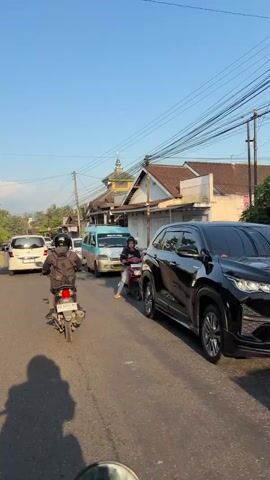 The video shows a street scene with vehicles and buildings under a clear sky. There is traffic congestion on the road, with cars and motorcycles. A light blue van is in the center of the frame, and a white car is behind it. A motorcycle is in the foreground, with its rider facing away from the camera. Another motorcycle, ridden by a woman wearing a hijab, is to the right of the frame. Buildings with tiled roofs line the street, and a mosque with a green dome is visible in the background. Power lines are strung across the sky. The overall impression is a busy street in a residential or commercial area.