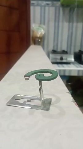The video shows a close-up of a lit mosquito coil on a metal stand, resting on a white surface with a patterned design. The coil is green and coiled into a spiral shape. A small amount of smoke is rising from the lit end of the coil. In the background, a blurred kitchen counter is visible with a stove and some items on it. The overall lighting suggests an indoor setting.