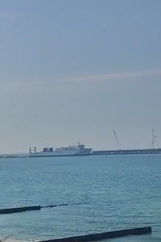 The video shows a blue sea with a ship in the distance to the left. There is also a long structure in the water with several cranes on it. In the foreground, there are several wooden breakwaters extending into the water. The sky is pale blue with some light clouds. The camera pans to the right, eventually showing only the ocean and the sky.