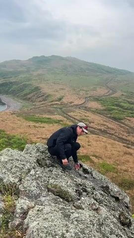 A man in a black jacket, pants, and cap is seen crouching on a rocky outcrop, then standing up and walking to the edge of the rock. He looks out at the view. The background shows a grassy, hilly landscape with a body of water on the left side of the frame. The sky is overcast.