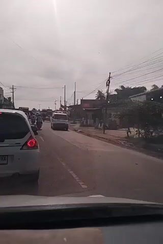 The video shows a white ambulance with red flashing lights driving on a road. The ambulance has the word "AMBULANCE" written on the side. The road is lined with trees and buildings. There are other vehicles on the road, including cars and trucks. The sky is cloudy. The video is shot from inside another vehicle, with the dashboard visible in the foreground.