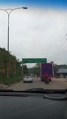 A shot from inside a vehicle looking out at a road with traffic. A green sign above the road reads "Bucaramanga, Centro de la Moda, Calzado y Joyeria". A person on a motorcycle wearing a red shirt is in the foreground, followed by a white car and a large purple truck. Trees and hills are visible on the side of the road. The sky is overcast.