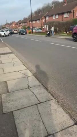 The video shows a residential street with houses on both sides. A black car drives along the road. On the right side of the road, a person is pushing a stroller. A sidewalk is visible on the left side of the road. The sky is overcast.