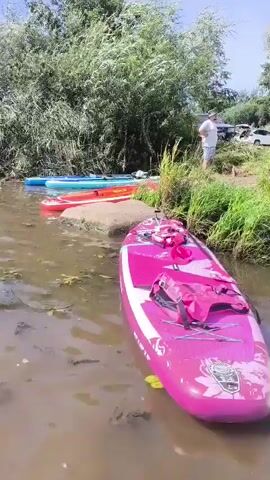 The video shows a group of people on the bank of a body of water. Several paddleboards are in the water near the shore. In the foreground, a pink paddleboard with a floral design is partially submerged. To its right, a white paddleboard with blue and red accents is visible. Further back, a green and red paddleboard and another blue and white paddleboard are present. In the background, several people are standing on the grassy bank. Some are talking, and one person is wearing a hat. There are trees and bushes behind the people. Cars are parked in the background. The water appears calm and shallow near the shore.