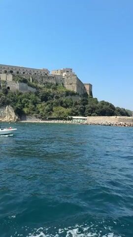 A small white boat with at least three people aboard navigates calm blue water near a rocky shore. A large, ancient-looking stone fortress with multiple buildings sits atop a verdant, tree-covered hill overlooking the water. A bright red lighthouse stands on a stone breakwater to the right of the fortress. The sky is clear and blue.